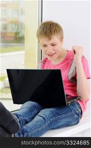 The joyful teenager with a computer sits on a window
