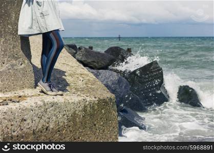 The legs of a young woman wearing a dress as she is standing on a rock by the coast with the waves crashing in