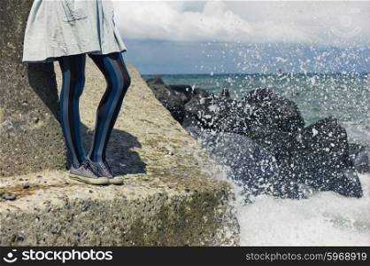 The legs of a young woman wearing a dress as she is standing on a rock by the coast with the waves crashing in