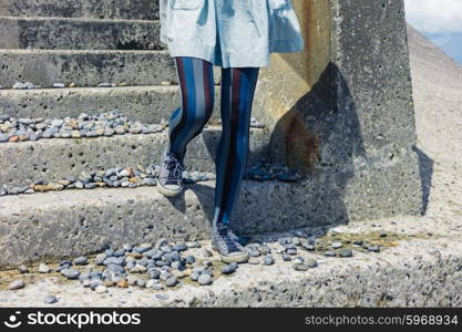 The legs of a young woman wearing a dress as she is standing on some stairs by the coast