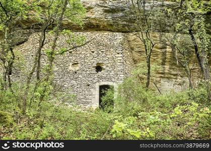 The main exterior wall of an abandoned structure of a house or a stable that is located underneath an overhanging cliff in the forests of the Luberon area in Provence, France.