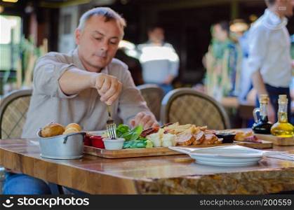 The man is eating breakfast in the restaurant.There are a variety and delicious breakfast on the table.he is picking olives with fork.