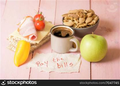 "The message "pay the bills" wrote on a piece of paper placed on a pink wooden table, near a small cup of coffee and a morning meal"
