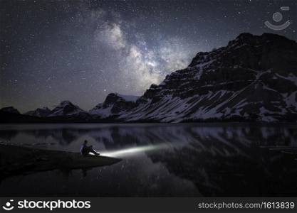 The Milky Way shining over Bow Lake and Crowfoot Mountain alongside the Icefields Parkway in Banff National Park while a stargazer illuminates the blue-green waters of the icy lake.