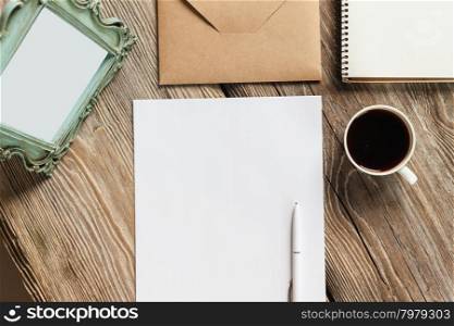 The mockup on wooden background with vintage old picture frame, pen, pencil, cup of coffee, white blank paper for writing, envelope and notebook