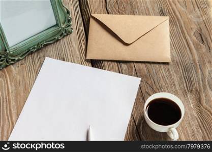 The mockup on wooden background with vintage old picture frame, pen, pencil, cup of coffee, white blank paper for writing, envelope and notebook