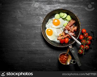 The morning Breakfast in a frying pan with a fork. Fried eggs with bacon, beans and cucumbers. On a black wooden background.. The morning breakfast in a frying pan