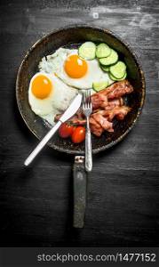 The morning Breakfast in a frying pan with a fork. Fried eggs with bacon, beans and cucumbers. On a black wooden background.. The morning Breakfast