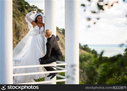 The newlyweds are walking in a beautiful picturesque gazebo, the girl is holding a veil with her hand, the guy is sitting on the railing