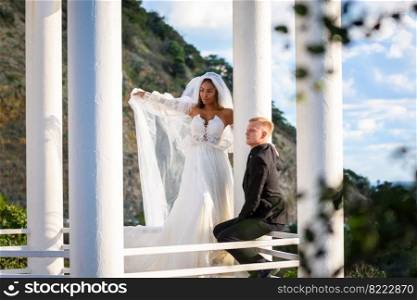 The newlyweds are walking in the gazebo, the girl is standing with her veil raised, the guy is sitting on the railing