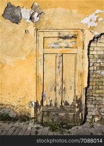 the old brick wall of the house from which part of the plaster has fallen off and destroyed, close-up of an old building during the period of its destruction, closed yellow wooden door. closed yellow wooden door