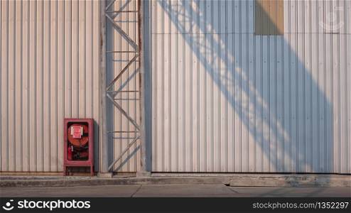 The old red emergency cabinet and rustic metal overhead signpost structure on concrete floor with sunlight and shadow on metal sheet wall surface of factory building, security in industrial concept
