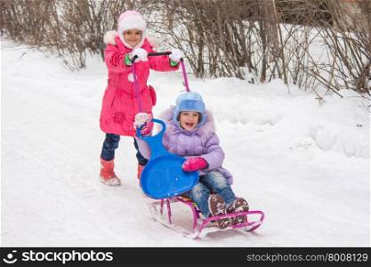 The older girl rolls the younger girl on a sled in the yard. Two girls girlfriends ride each other on a sled in the snowy winter weather