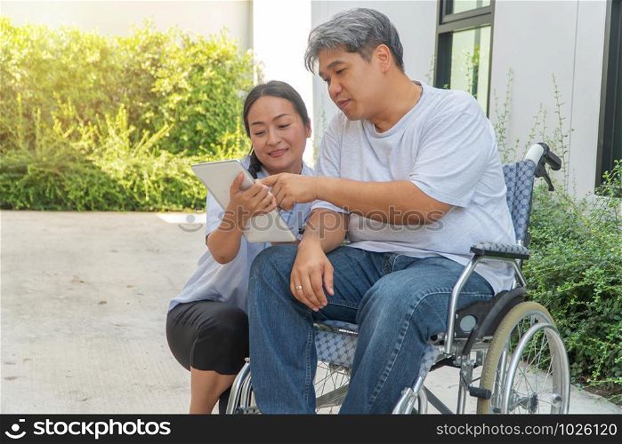 The patient caused by a car accident sitting in a wheelchair and his wife is beside And holding a tablet Let him look to relax while strolling in the garden. Concept of being by side and encouraging.