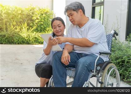 The patient caused by a car accident sitting in a wheelchair and his wife is beside And holding a tablet Let him look to relax while strolling in the garden. Concept of being by side and encouraging.