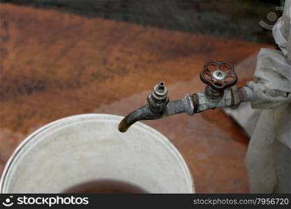 The picture of the old rusty water tap, crocked film. Under the tap is a plastic bucket with dirty water.