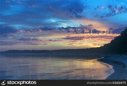 The picturesque night sky with colorful clouds at sunset reflecting in the golden surface of the water. Midnight on the forested shore of Lake Onega in the White Nights season. Republic of Karelia, Russia.