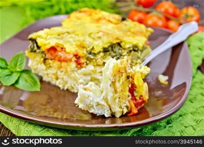 The pie of potatoes, cheese, tomato and spinach, filled egg with milk in a plate with a fork on a green napkin on a wooden boards background