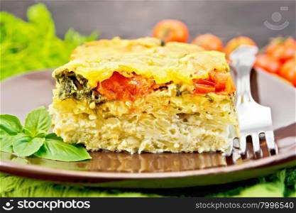The pie of potatoes, cheese, tomato and spinach, filled egg with milk in a plate with a fork, a napkin on a dark wooden board