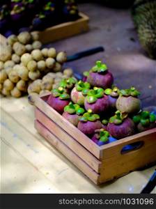 The pile of mangosteen and wollongong are in a small wooden crate. Put on a light yellow surface Inside the outdoor market, selective focus.
