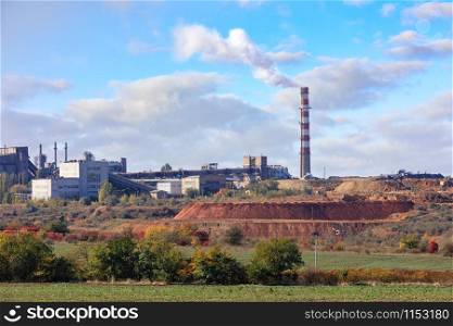 The pipe of the cement plant rises above the quarry of clay and limestone against the background of a slightly cloudy blue summer sky.. Cement factory on the background of a quarry of limestone and clay on a summer day.