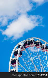 The pods of a Ferris wheel set infront of a backdrop of blue sky and white clouds, viewed from below.