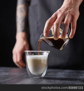 The process of making coffee with milk. A man&rsquo;s hand pours coffee into a cup of milk on a black wooden table.. A man&rsquo;s hand pours coffee into a cup of milk on a black wooden table. Cappuccino preparation.