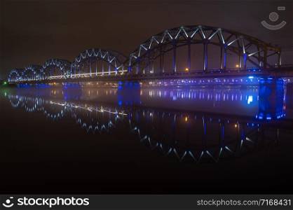 The Railway Bridge in Riga at foggy winter night with beautiful reflections in still water of Daugava river. Train crossing the bridge. Long exposure
