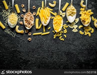 The range of different types of dry pasta in spoons. On black rustic background. The range of different types of dry pasta in spoons.