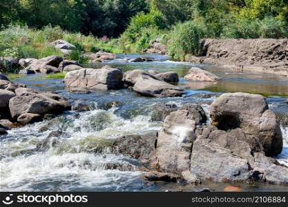 The rapid current of the clear waters of a forest stream makes its way through the large stone boulders of the rapids against the backdrop of a thicket and a blue sky.. Beautiful stone rapids on the way of a fast flow of a small forest stream on a sunny summer day.