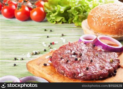 The raw ingredients for the homemade burger on green wooden background. Beef cutlet, cheese, tomato, salad, onion, wheat bun, spice. The ingredients for the burger on green wooden background