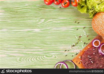 The raw ingredients for the homemade burger on green wooden background. Beef cutlet, cheese, tomato, salad, onion, wheat bun, spice. Top view, flat lay, mockup with copy space for text. The ingredients for the burger on green wooden background