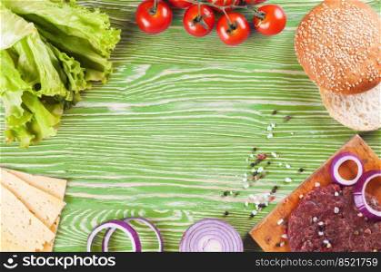 The raw ingredients for the homemade burger on green wooden background. Beef cutlet, cheese, tomato, salad, onion, wheat bun, spice. Top view, flat lay, mockup with copy space for text. The ingredients for the burger on green wooden background