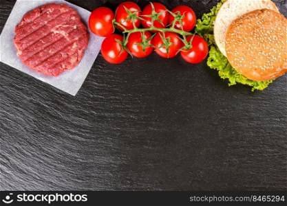 The raw ingredients for the homemade burger. Raw coutlet, tomatoes, cheese, salad, wheat bun on black slate background on the right side with copy space for text. Top view. Flat lay.. The raw ingredients for the homemade burger on black slate background. Top view.