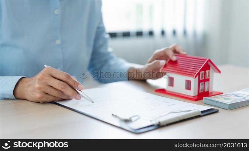 The real estate concept The man in blue shirt, real estate agent, holding a pen in his right hand and a house model in his left hand and reading the contract before handing it to his customer.