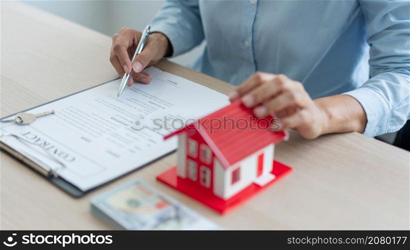 The real estate concept The man in blue shirt, real estate agent, holding a pen in his right hand and a house model in his left hand and reading the contract before handing it to his customer.