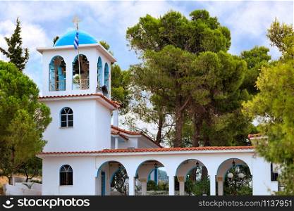 The ritual bell tower of the Orthodox Church of Ekklisia Agios Ioannis in Loutraki surrounded by green Mediterranean spruces, August 15, 2019, Greece.. Exterior of the traditional white-blue Greek bell tower of a Christian Orthodox temple in Loutraki, Greece.