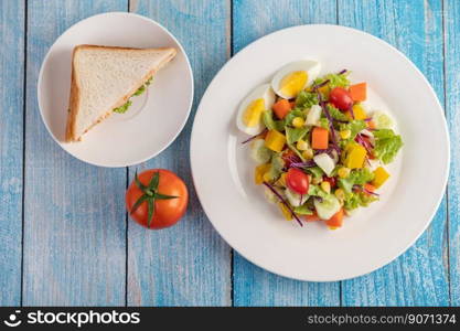 The salad is on a white plate, with a sandwich and tomatoes on the blue wooden floor.