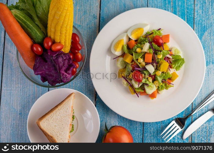 The salad is on a white plate, with a sandwich and tomatoes on the blue wooden floor.