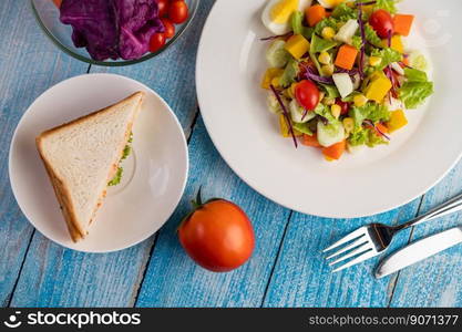 The salad is on a white plate, with a sandwich and tomatoes on the blue wooden floor.