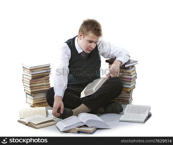 The schoolboy in a school uniform sits on a floor, near to packs of books, with the opened book in hands