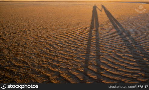 The shadow of a very long couple holding hand on a sand beach surface.