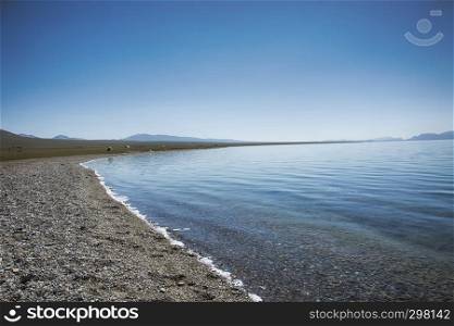 The shore of the lake in the early morning, quiet and windless weather, the shore with shallow round pebbles, a clear blue sky. Son-kul lake. National park of the Kyrgyzstan.