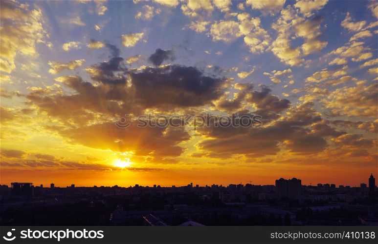 The silhouette of a sleeping city against the background of a fiery reddish sunrise, the rays of which are dispersed by the clouds. Fiery reddish sunrise over a sleeping city