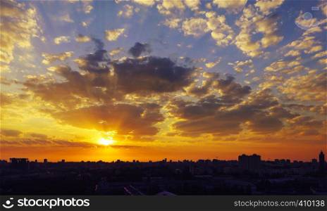 The silhouette of a sleeping city against the background of a fiery reddish sunrise, the rays of which are dispersed by the clouds. Fiery reddish sunrise over a sleeping city