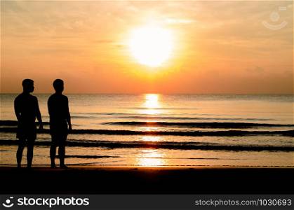 The silhouette photo of the gay couple standing together on the beach enjoy sunrise moment with the reflection on the sea in summer season for vacation trip.