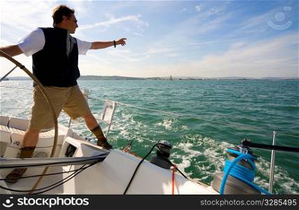 The skipper of a yacht points towards the horizon while keeping one hand on the boat&acute;s wheel