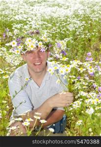 The smiling man in a wreath from wild flowers in the field of camomiles with a bouquet
