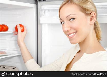 The smiling woman takes a tomato from a refrigerator. Storage of products