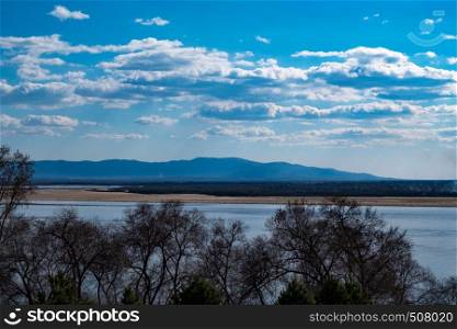 The spring Park is illuminated by the bright sun. View of a large and powerful river. On the bright blue sky beautiful white clouds.. View of the Amur river against the blue sky with white beautiful clouds. Bright spring sun. Russia, Khabarovsk.
